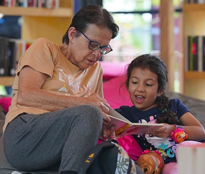 Mujer y niña leen juntas un libro en una biblioteca de Comfama
