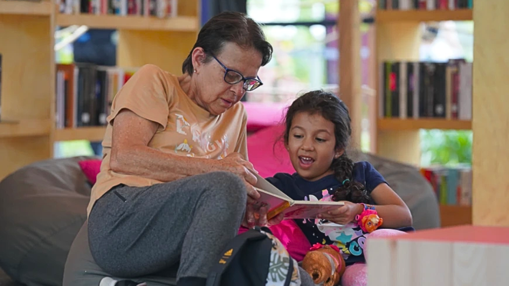 Mujer y niña leen juntas un libro en una biblioteca de Comfama